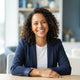 A friendly, diverse woman smiling confidently in a professional headshot against a soft-focus blue and white background.